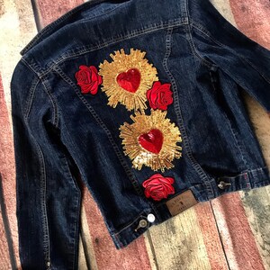 May include: A dark blue denim jacket with three red roses and two gold heart-shaped patches with red sequins. The patches have a gold sunburst design around the hearts.