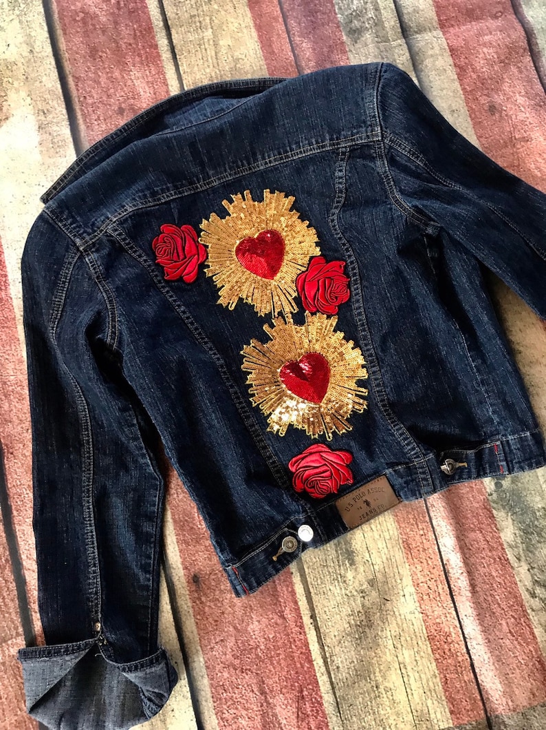 May include: A dark blue denim jacket with three red roses and two gold heart-shaped patches with red sequins. The patches have a gold sunburst design around the hearts.
