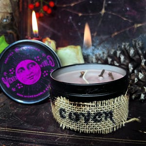 Könnte beinhalten: Eine schwarze Blechkerze mit einem Jute-Etikett, auf dem "Giver" steht. Die Kerze ist angezündet und hat ein rosa Wachs. Die Dose hat ein schwarzes und violettes Etikett mit dem Text "Wicked World Oddities" und einem Mond mit einem Gesicht.