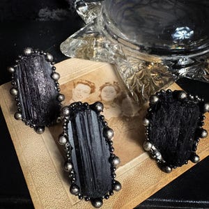 May include: Three rings featuring raw, vertically striated black tourmaline crystals set in dark, oxidized metal bands. Each stone is surrounded by a decorative border of small, round metal beads, creating a textured, rugged appearance for these jewelry pieces.