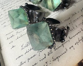 May include: Four rings featuring large, translucent, pale green cubic crystals set in dark, oxidized metal bands. Each ring is accented with small, spherical metal beads along the edges of the setting, framing the geometric, raw-cut stone.
