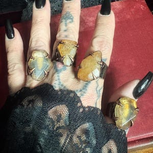 May include: Four rings featuring raw, translucent yellow and amber-toned gemstones set in dark, textured metal bands with claw-like prongs. Each ring displays a unique, irregular stone shape, showcasing natural inclusions and a polished surface finish.