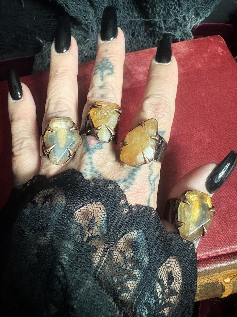 May include: Four rings featuring raw, translucent yellow and amber-toned gemstones set in dark, textured metal bands with claw-like prongs. Each ring displays a unique, irregular stone shape, showcasing natural inclusions and a polished surface finish.