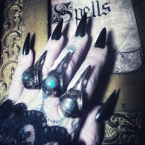 May include: A hand wearing several black rings with blue gemstones. The rings are shaped like bird skulls. The hand is wearing long black nails and is resting on a vintage book with the word "Spells" written on it.