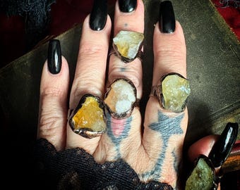 May include: Five rings featuring raw, uncut gemstones set in dark, oxidized metal bands. The stones display various shades of yellow, amber, and pale green. Each ring has a unique, irregular shape, showcasing the natural texture of the crystals.