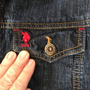 May include: Close-up of a dark blue denim jacket with a red and white embroidered polo player logo on the pocket. The pocket has a gold button with the text "US POLO ASSOC. JEANS CO."