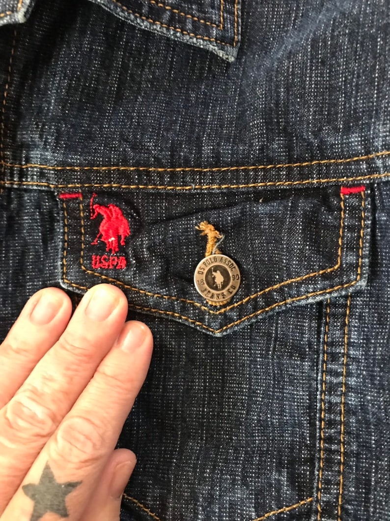 May include: Close-up of a dark blue denim jacket with a red and white embroidered polo player logo on the pocket. The pocket has a gold button with the text "US POLO ASSOC. JEANS CO."