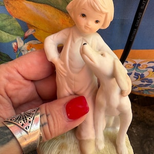 Peut inclure: Figurine en porcelaine repr&eacute;sentant un enfant et un chien. L'enfant, aux cheveux blonds courts, porte une tenue blanche et a une main sur la hanche. Le chien blanc se dresse sur ses pattes arri&egrave;re, regardant l'enfant.