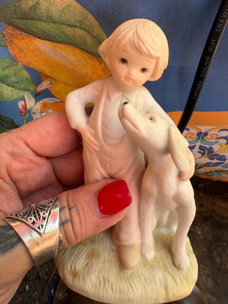 Peut inclure: Figurine en porcelaine repr&eacute;sentant un enfant et un chien. L'enfant, aux cheveux blonds courts, porte une tenue blanche et a une main sur la hanche. Le chien blanc se dresse sur ses pattes arri&egrave;re, regardant l'enfant.