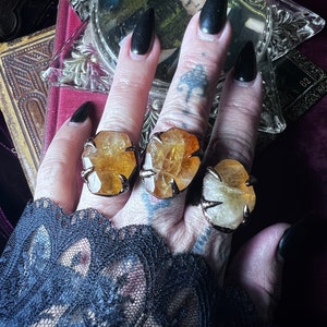 May include: Three gold rings with large, claw-shaped settings featuring yellow and orange citrine stones. The rings are worn on the fingers of a hand with black nail polish.