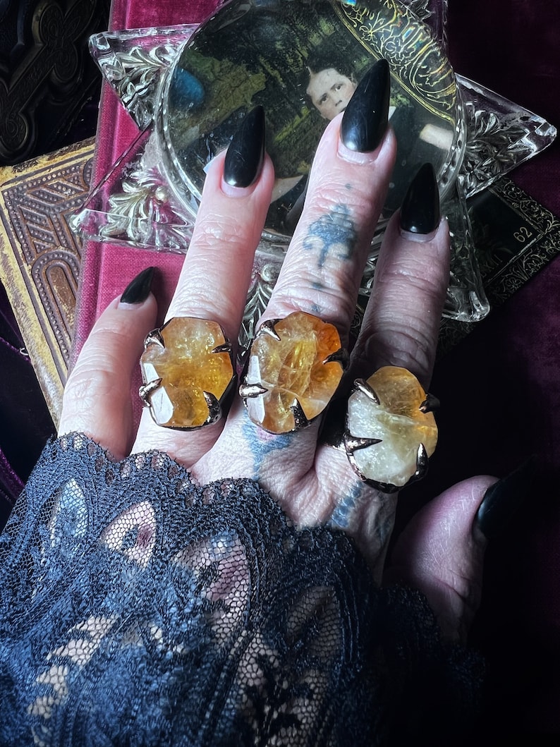 May include: Three rings featuring large, raw, golden-yellow citrine gemstones set in dark, metallic claw-style prongs. Each stone displays a faceted, irregular shape with natural inclusions, secured by four metal prongs on a dark band.