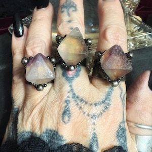 May include: Three rings featuring faceted, pyramid-shaped crystals set in dark, textured metal bands. Each crystal displays translucent hues of pale purple, yellow, and clear tones, accented by small, round metallic beads at the base of the settings.