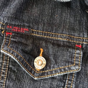 May include: Close-up of a dark blue denim jacket with a brown pocket and a silver button with the text "U.S. POLO ASSOC. JEANS CO."
