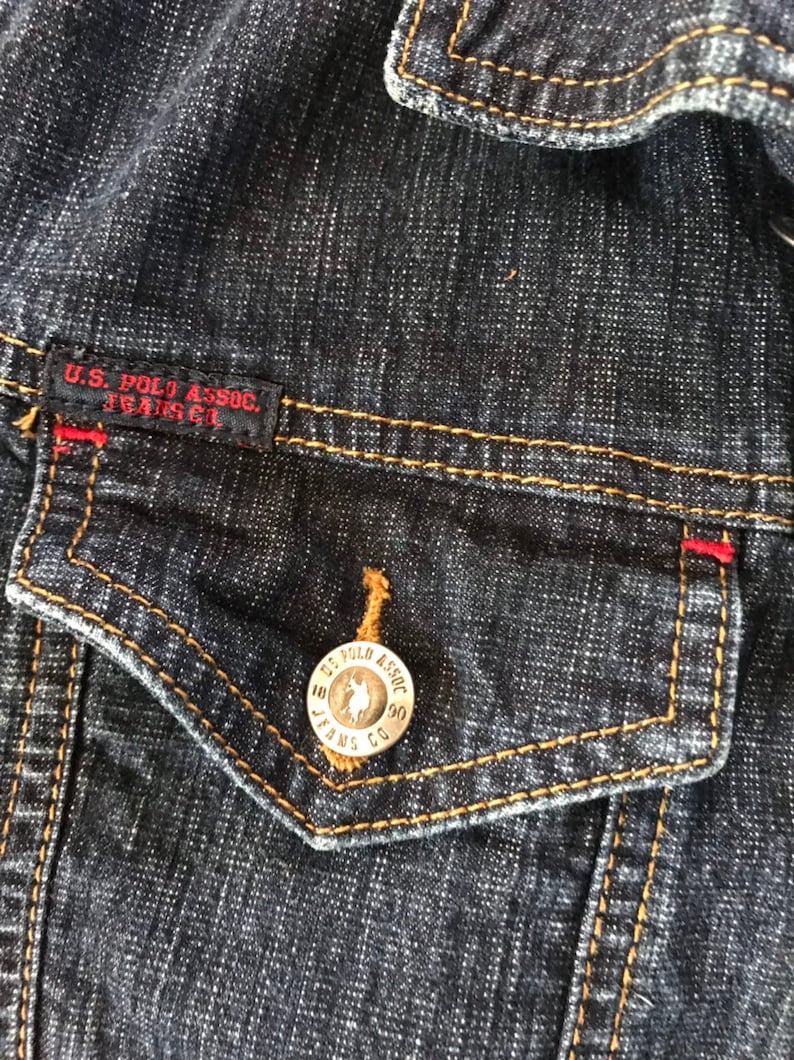 May include: Close-up of a dark blue denim jacket with a brown pocket and a silver button with the text "U.S. POLO ASSOC. JEANS CO."