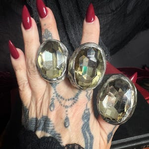 May include: Three rings featuring large, oval-shaped, faceted light yellow stones set in textured, metallic silver-toned bands. Each ring displays a prominent, reflective gemstone with multiple facets, secured within a thick, irregular metal setting.