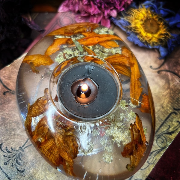 May include: A clear glass candle holder with dried flowers and a lit candle inside. The holder is shaped like a rounded rectangle and has a black metal base.