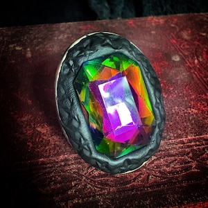 May include: A ring featuring a large, rectangular, multi-faceted iridescent gemstone that displays vibrant rainbow colors including purple, green, and yellow. The stone is set within a dark, textured, matte black frame with a repeating pattern.