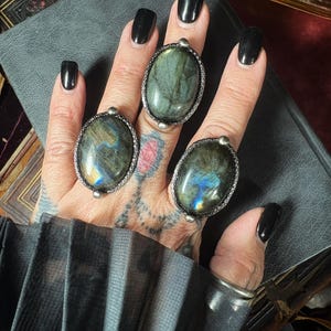 May include: Three oval rings featuring large, polished labradorite gemstones with iridescent blue and gold flashes. Each stone is set in a textured, dark silver-toned metal bezel with small decorative spheres at the top and bottom of the setting.