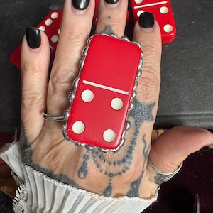 May include: A statement ring featuring a red domino tile with white pips, set in a scalloped silver-toned metal frame. The domino displays a blank top section and four white dots on the bottom half, secured by a simple silver-toned metal band.