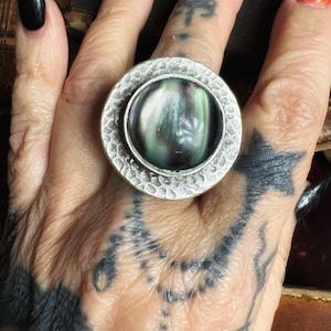 May include: A circular ring featuring a wide, hammered silver-tone band and a central, domed cabochon stone. The stone displays a dark, iridescent surface with swirling hues of green, purple, and black, resembling the appearance of an aurora.