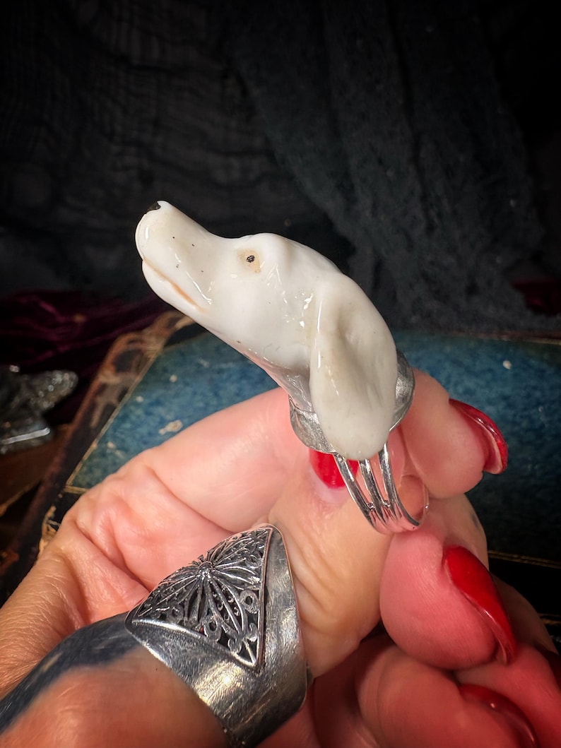 Peut inclure: Bague t&ecirc;te de chien en c&eacute;ramique blanche avec une monture argent&eacute;e. La t&ecirc;te de chien est d&eacute;taill&eacute;e avec un &oelig;il noir et une bouche l&eacute;g&egrave;rement ouverte. La bague est port&eacute;e sur un doigt vernis rouge.