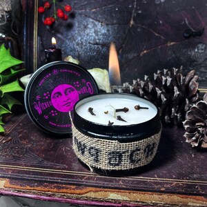 Könnte beinhalten: Eine schwarze Kerzen-Dose mit einem Jute-Etikett, auf dem "WITCH" steht, mit einer weißen Kerze darin. Die Kerze ist angezündet und hat ein paar Nelken oben drauf. Die Dose hat ein Etikett, auf dem "Oddities & Curiosities of the Wicked World" steht.