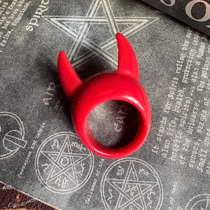 May include: A bright red ring shaped like devil horns. The ring is set against a backdrop of a book and a dark gray surface with white pentagram and text details. The ring is a bold, eye-catching accessory.