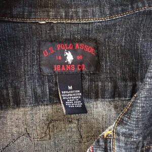 May include: A black and red label with the text "U.S. POLO ASSOC. JEANS CO." and a small image of a polo player on a horse. The label is attached to a dark blue denim shirt. The label also includes the text "M 88% COTTON 10%POLYESTER 20%SPANDEX MADE IN HONG KONG SEE REVERSE FOR CARE."