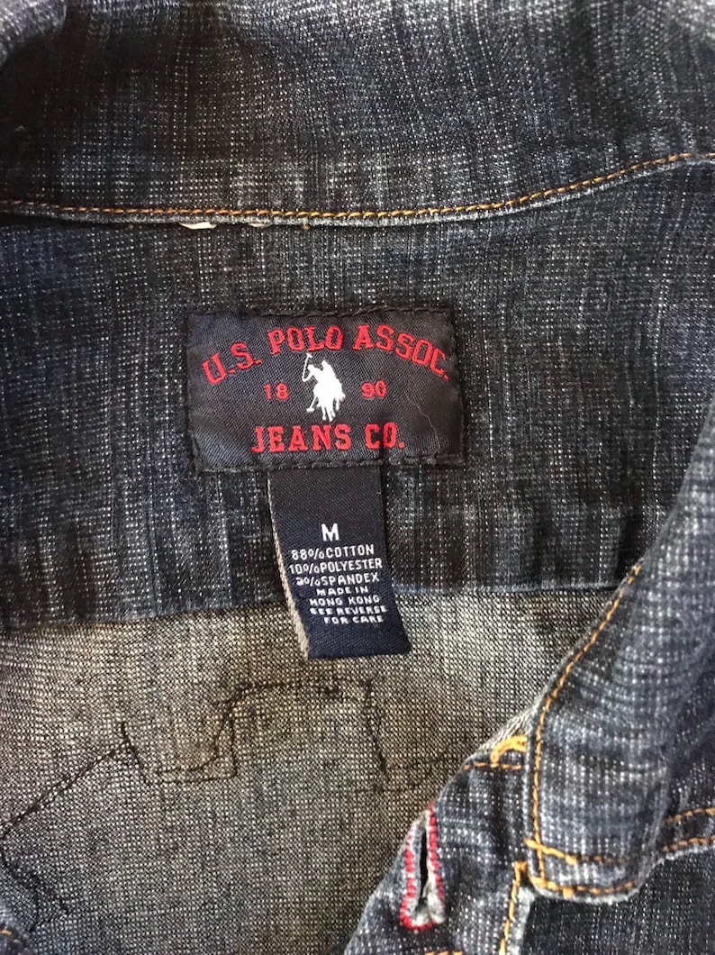 May include: A black and red label with the text "U.S. POLO ASSOC. JEANS CO." and a small image of a polo player on a horse. The label is attached to a dark blue denim shirt. The label also includes the text "M 88% COTTON 10%POLYESTER 20%SPANDEX MADE IN HONG KONG SEE REVERSE FOR CARE."