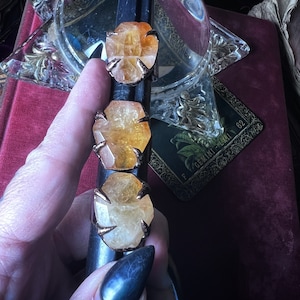 May include: Three rings featuring raw, faceted orange-yellow citrine stones set in dark metal prongs. The stones are arranged vertically on a black cylindrical display stand, showcasing their natural, irregular shapes and translucent, warm-toned crystalline structure.
