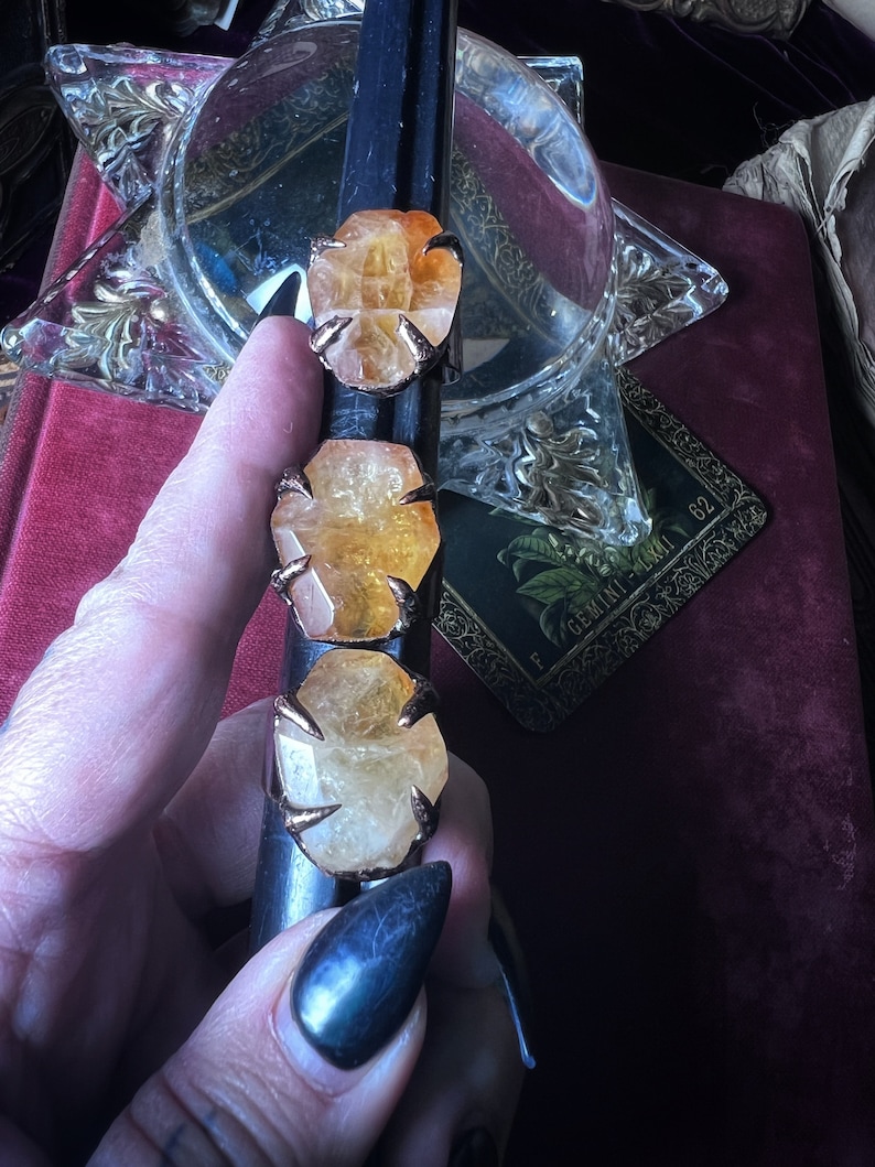 May include: Three rings featuring raw, faceted orange-yellow citrine stones set in dark metal prongs. The stones are arranged vertically on a black cylindrical display stand, showcasing their natural, irregular shapes and translucent, warm-toned crystalline structure.