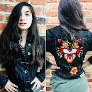 May include: A black denim jacket with a leopard print design and an embroidered tiger face on the back. The tiger has orange, yellow, and red fur.