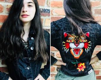 May include: A black denim jacket with a leopard print design and an embroidered tiger face on the back. The tiger has orange, yellow, and red fur.