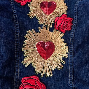 May include: A denim jacket with three red rose patches and two gold sequin hearts with red centers. The hearts are surrounded by gold sequin rays.