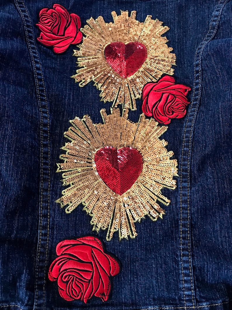 May include: A denim jacket with three red rose patches and two gold sequin hearts with red centers. The hearts are surrounded by gold sequin rays.