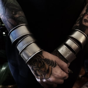 May include: A set of silver-toned metal cuff bracelets worn on both wrists. The cuffs feature a mix of smooth, polished surfaces and one textured band with an intricate, embossed pattern. The bracelets are wide and stackable in design.