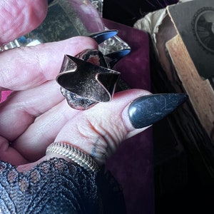 May include: A metallic ring featuring a wide, open-back band with a dark, oxidized finish and a textured, reddish-brown center stone. The ring has a prominent, angular design with sharp edges and a visible gap in the band, showcasing a raw, industrial aesthetic.