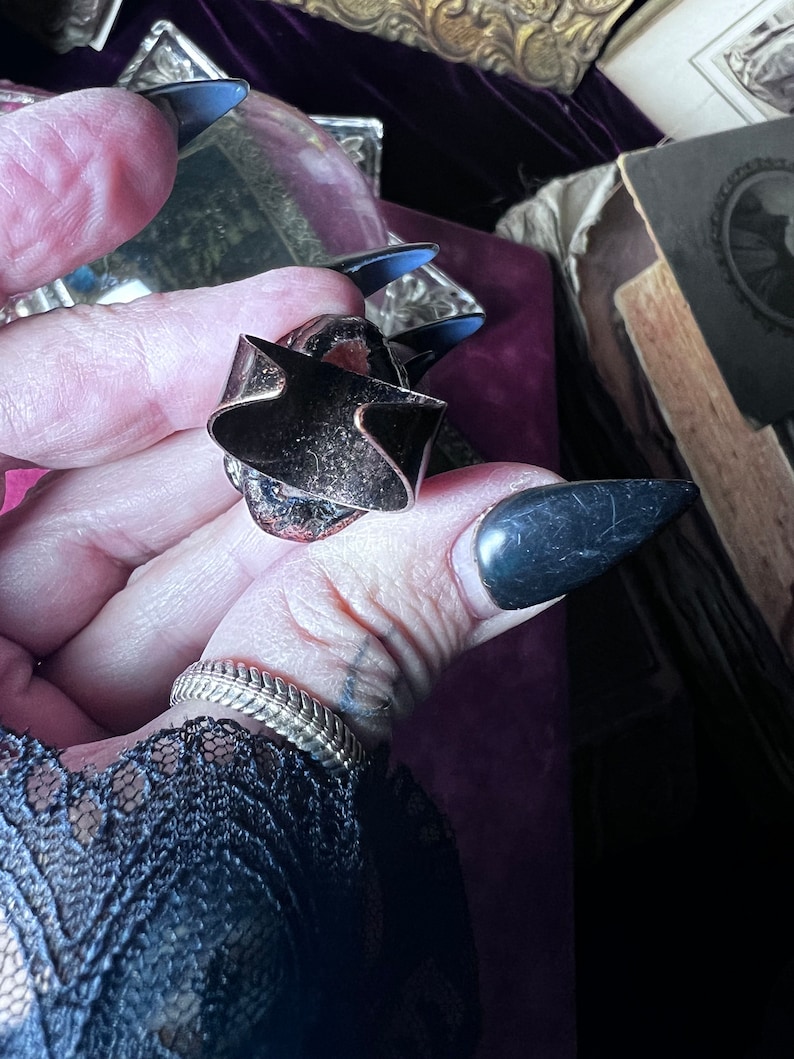 May include: A metallic ring featuring a wide, open-back band with a dark, oxidized finish and a textured, reddish-brown center stone. The ring has a prominent, angular design with sharp edges and a visible gap in the band, showcasing a raw, industrial aesthetic.