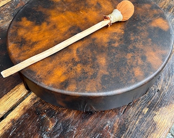 May include: A round frame drum with a mottled brown and black animal hide head, featuring a wooden mallet with a tan fabric-covered head and a light-colored wooden handle resting diagonally across the surface of the drum.