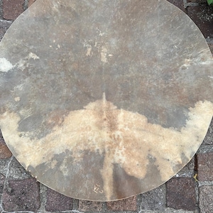 Pode incluir: Uma pele de tambor circular e plana com uma superfície de couro natural manchada, apresentando tons variados de bege, castanho e castanho escuro. A textura parece orgânica e irregular, com uma mancha clara distinta no centro e marcas visíveis.