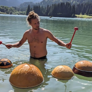 Pode incluir: Um homem toca um conjunto de quatro instrumentos de percussão de madeira flutuando em um lago. Os instrumentos são redondos e têm um acabamento de madeira natural. O homem está sem camisa e usa uma sunga preta. O lago é cercado por montanhas e árvores.