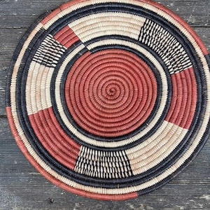 May include: A circular woven basket with a spiral pattern. The basket has concentric rings in red, black, white, and beige. It is made from natural fibres and has a rustic, handmade look.