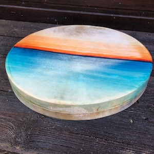 May include: A round, hand-painted drum with a blue, green, and orange sunset design. The drum is made of wood and has a natural finish.