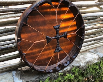 May include: A round frame drum with a dark brown wooden rim and a translucent amber-colored drumhead. The drumhead is tensioned by thin brown cords that meet in the center, where they are wrapped in black material to form a cross shape.