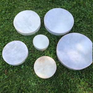 May include: Five round, white, leather-covered drums of varying sizes. The drums are arranged in a semi-circle on a green grassy surface.