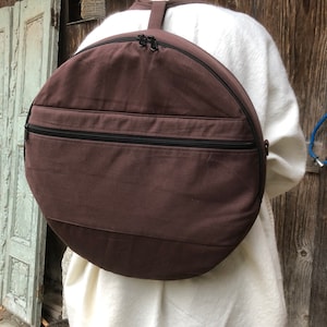 Può includere: Uno zaino in tela marrone con una grande apertura circolare e due tasche con cerniera. Lo zaino è progettato per trasportare un tamburo a cornice.