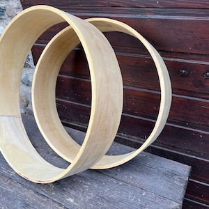 Op de afbeelding: Twee onafgewerkte houten hoepels, mogelijk voor knutselen of decoratie, worden getoond tegen een donkere houten achtergrond. De hoepels zijn licht van kleur, met een glad, gebogen ontwerp. Ze liggen op een verweerd houten oppervlak.