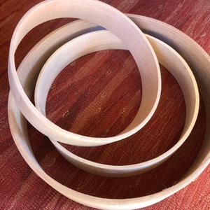 May include: Three wooden drum hoops of different sizes, nested together. The hoops are made of light-colored wood and have a smooth, polished finish.