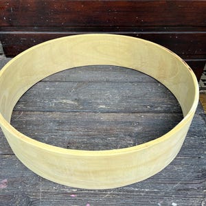 Könnte beinhalten: Ein helles, unfertiges Holzstück in Form eines Kreises, vermutlich ein Trommelreifen. Die Holzmaserung ist sichtbar. Der Reifen liegt auf einer verwitterten Holzoberfläche. Die Kanten sind abgerundet.