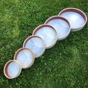 May include: A set of six round, wooden frame drums of varying sizes. The drums are arranged in a row from largest to smallest, with the largest drum at the top of the image. The drums are all white with a brown rim.